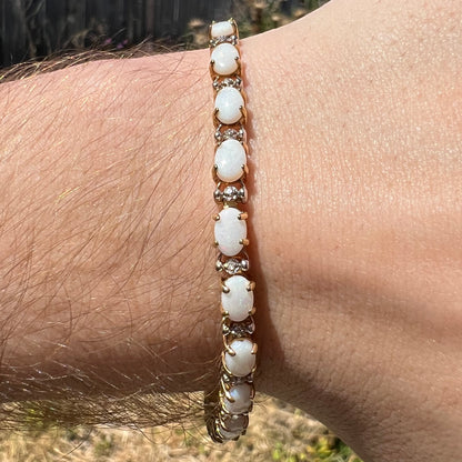 Close-up of an oval cut Australian opal and diamond tennis bracelet in gold, worn around a wrist in natural light.