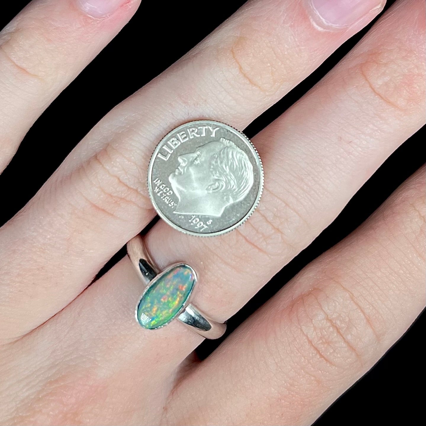 A sterling silver oval cut Lightning Ridge dark opal ring worn on a hand next to a US dime coin for scale.