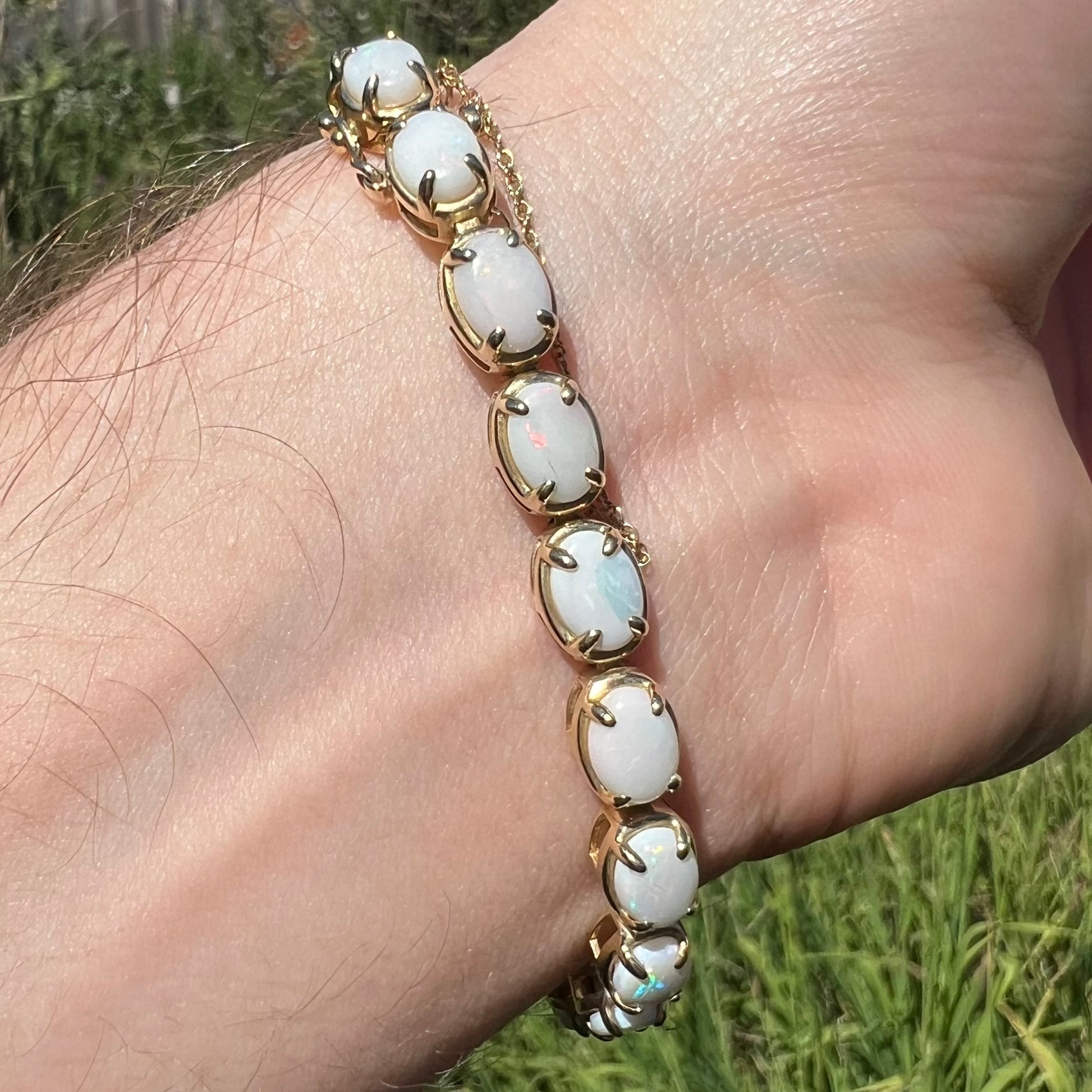 Underside of wrist wearing a yellow gold tennis bracelet prong-set with oval cut natural opals in sunlight.