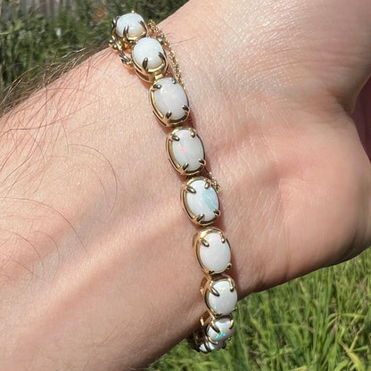 Underside of wrist wearing a yellow gold tennis bracelet prong-set with oval cut natural opals in sunlight.