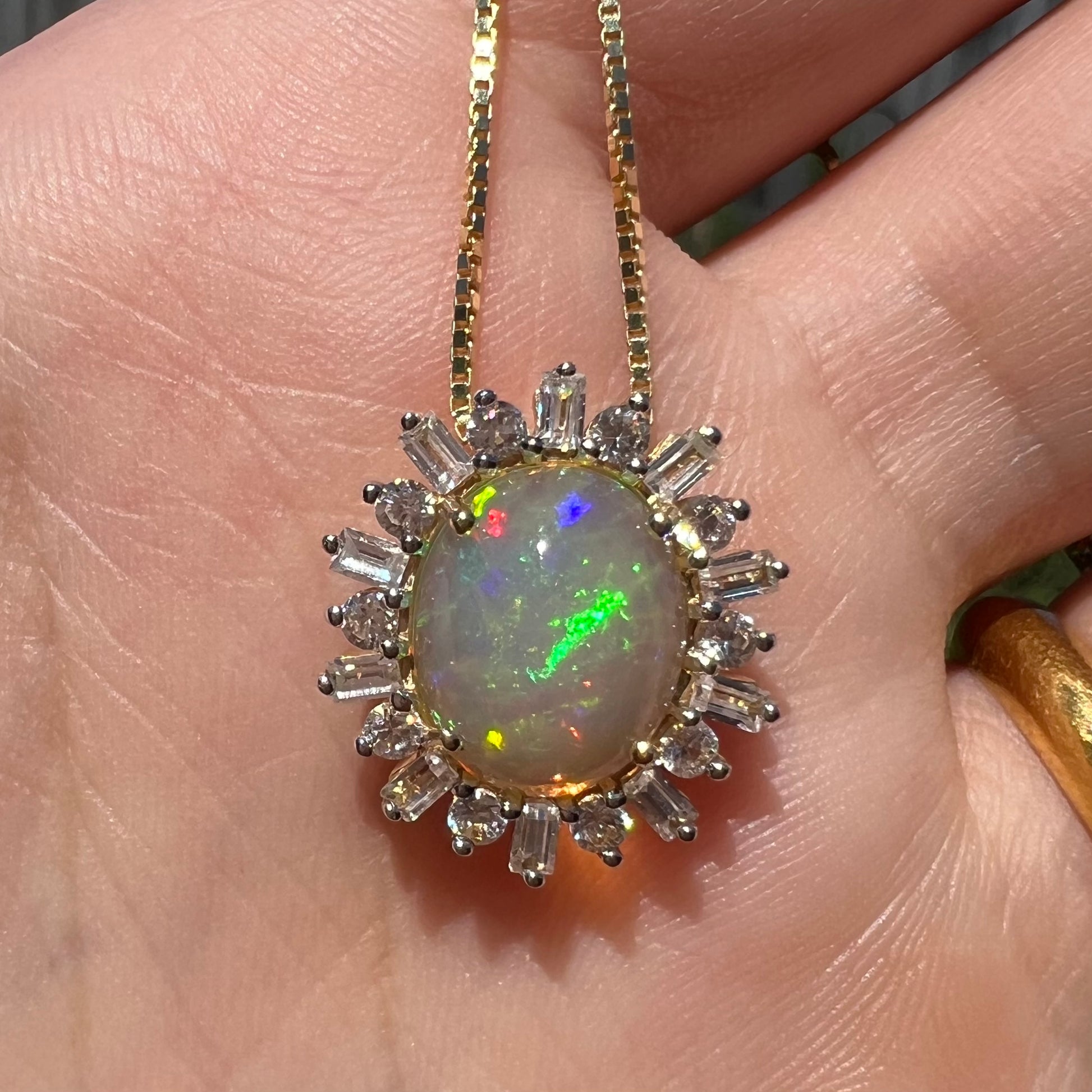Close-up of oval cut fire opal pendant with spiked halo design held in hand under natural light.  The opal has a green flash.