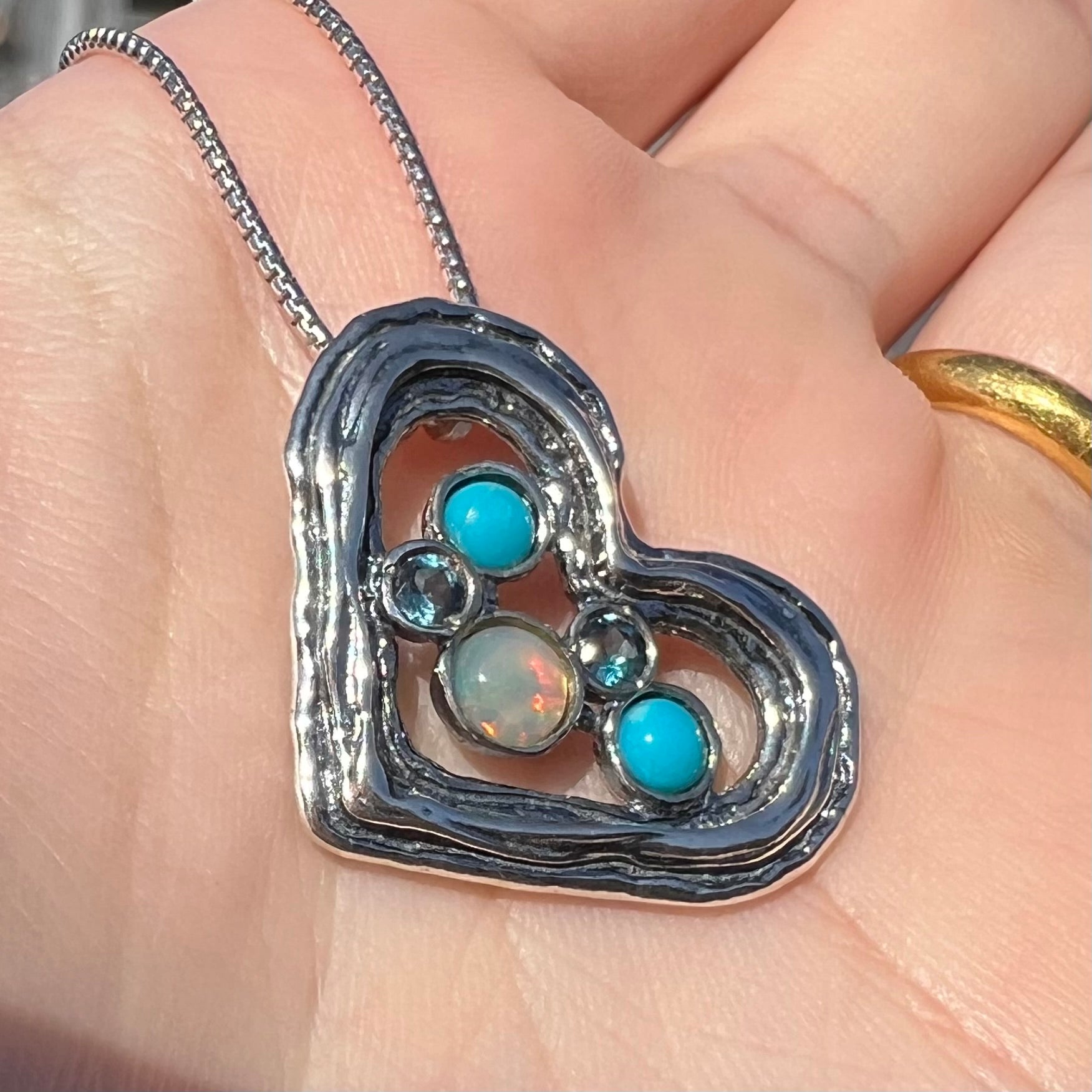 A sterling silver heart necklace set with opal, turquoise, and blue topaz shown close-up in sunlight at an angle.