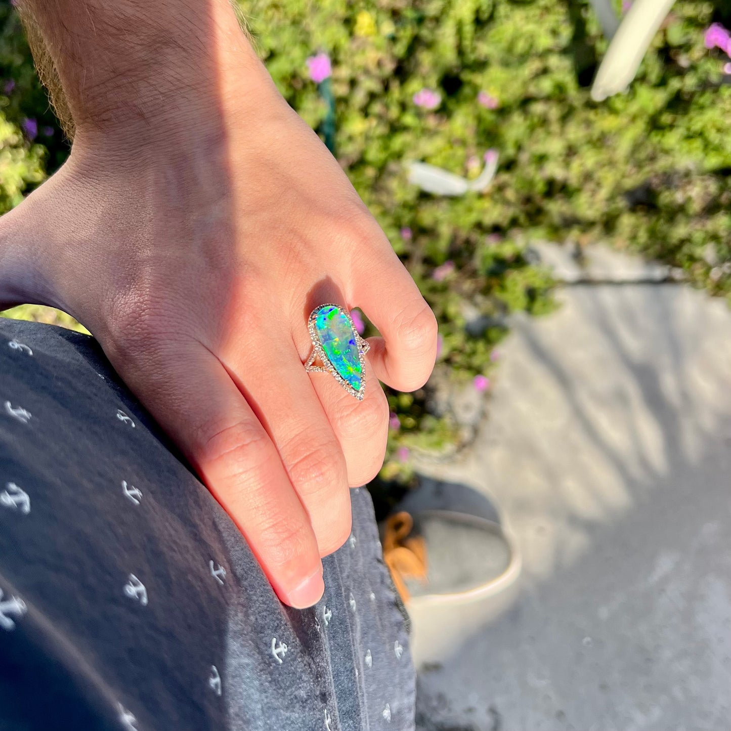 A hand resting on a person's hip while wearing a pear-shaped boulder opal and diamond ring in the sunlight against a blurred outdoor background.