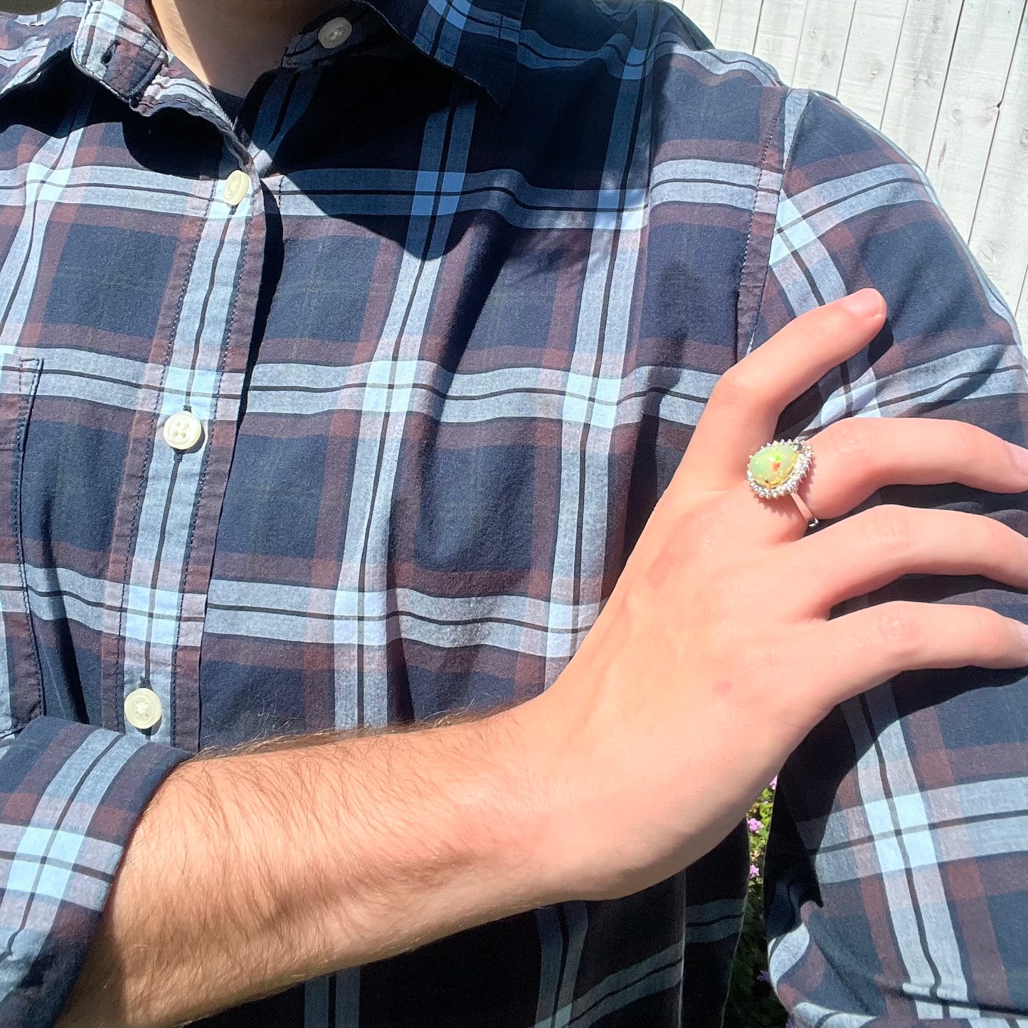 Person resting hand on shoulder in sunlight while wearing a white gold pear shaped fire opal and diamond ring with a blue plaid shirt.