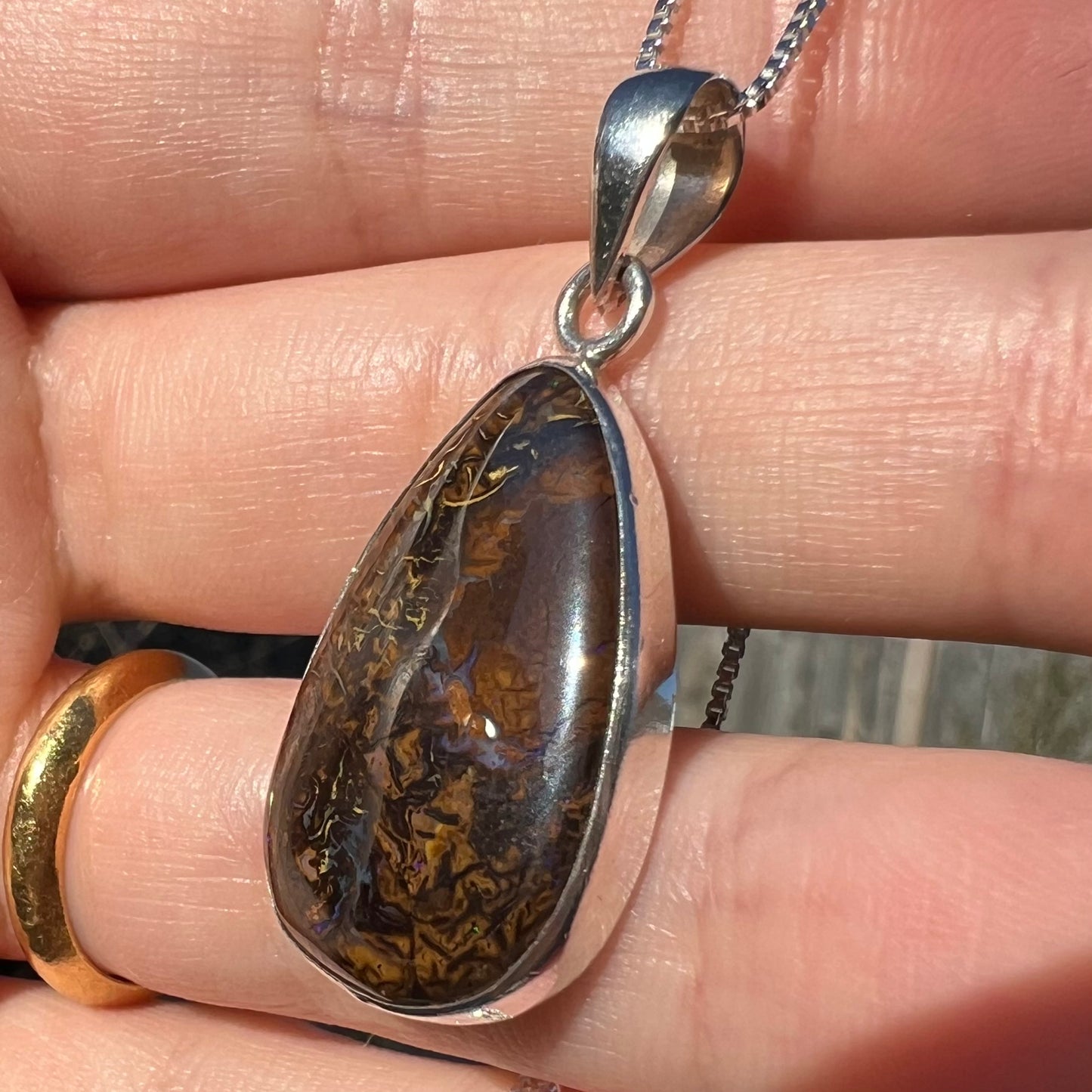 Angled close-up of a pear shaped Koroit boulder opal pendant in sterling silver shown in a hand in sunlight.