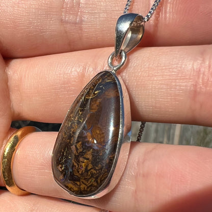 Angled close-up of a pear shaped Koroit boulder opal pendant in sterling silver shown in a hand in sunlight.