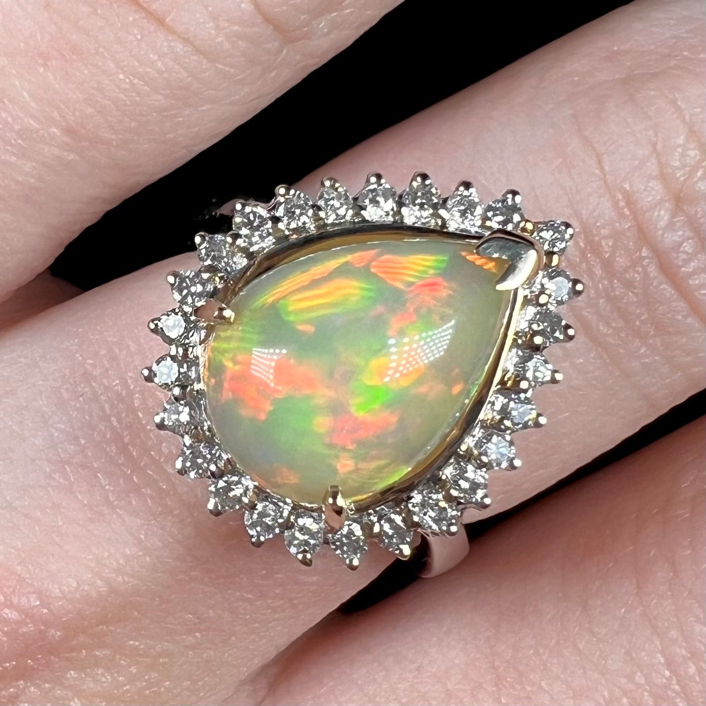 Close-up of a pear shaped fire opal and diamond halo ring with soft red and green play of color, worn on finger.