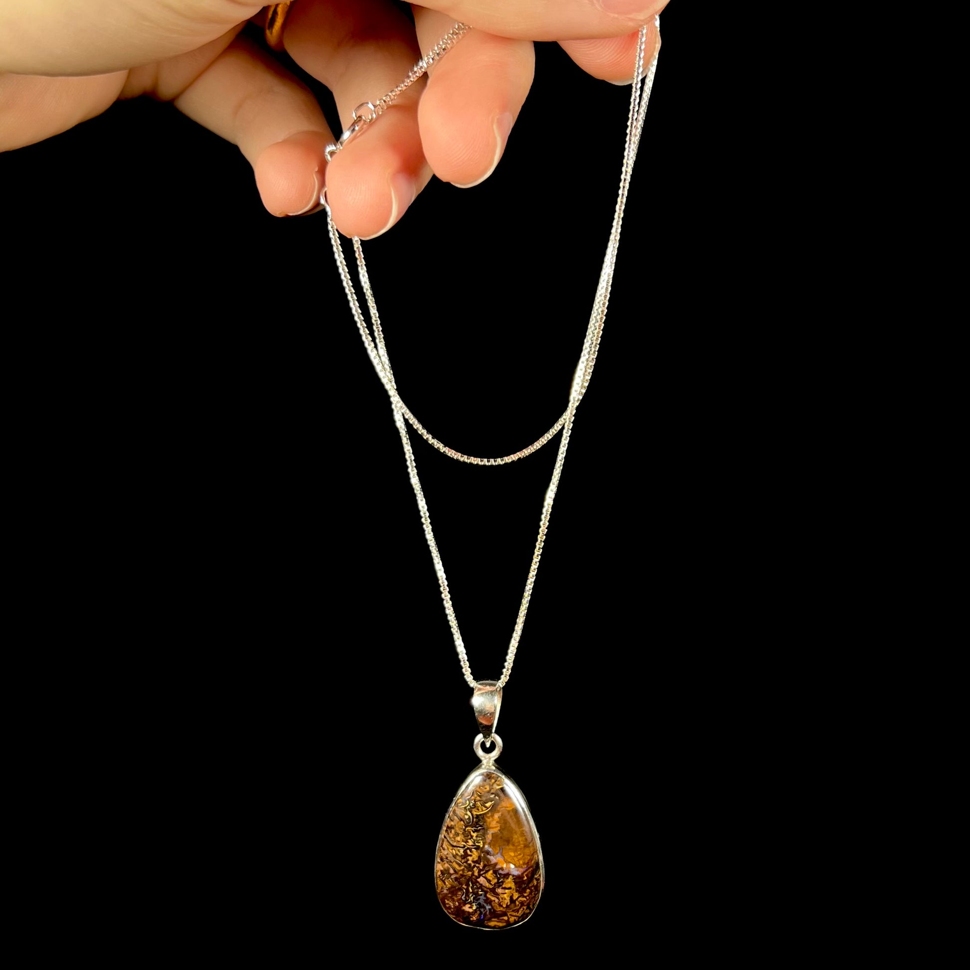 A pear shaped Koroit boulder opal necklace in sterling silver shown dangled by its chain by a hand.