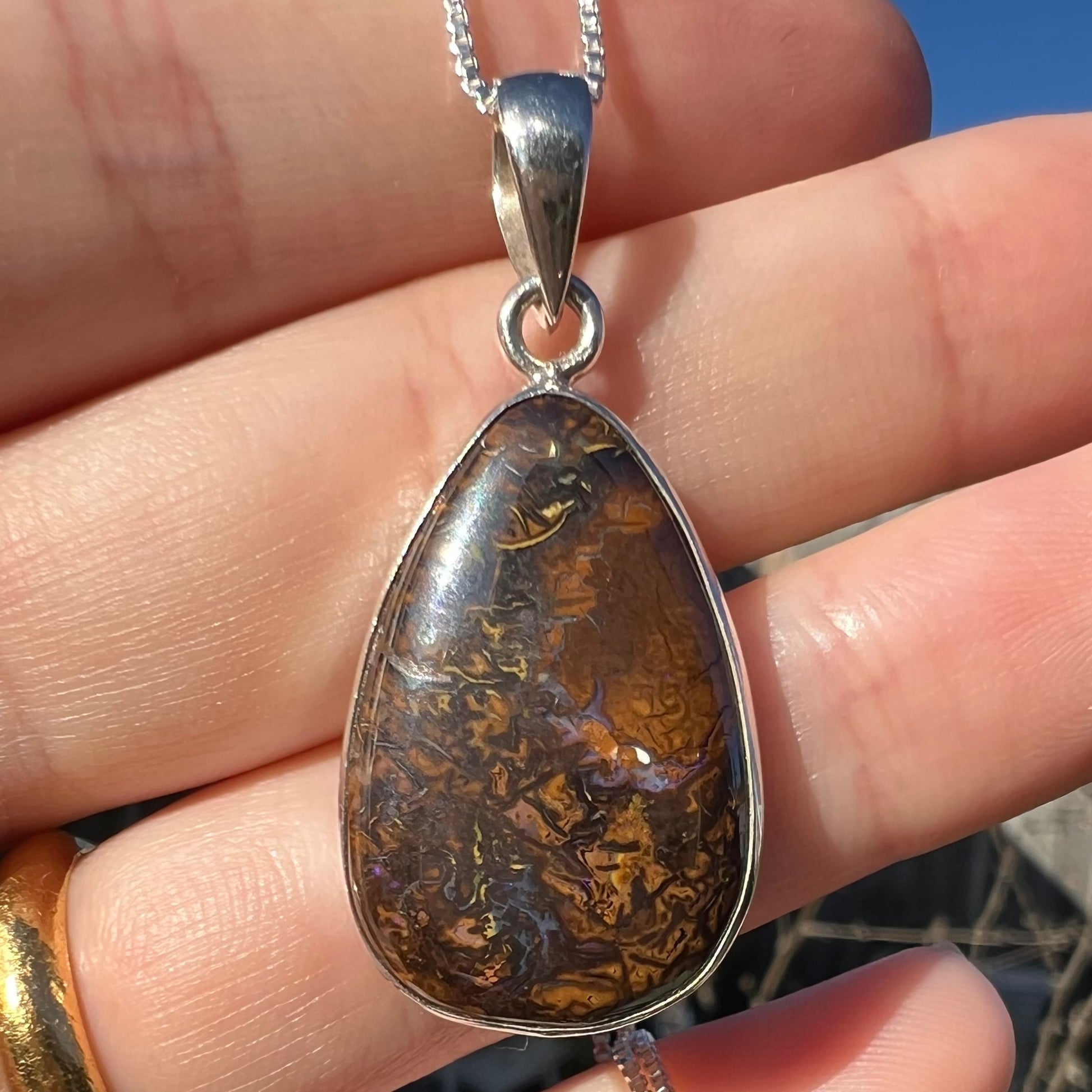 A pear shaped Koroit boulder opal pendant in sterling silver shown close-up in a hand in sunlight.