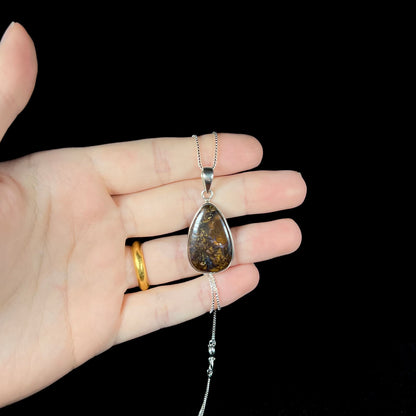 A pear shaped Koroit boulder opal necklace in sterling silver shown held in a hand against a black background.