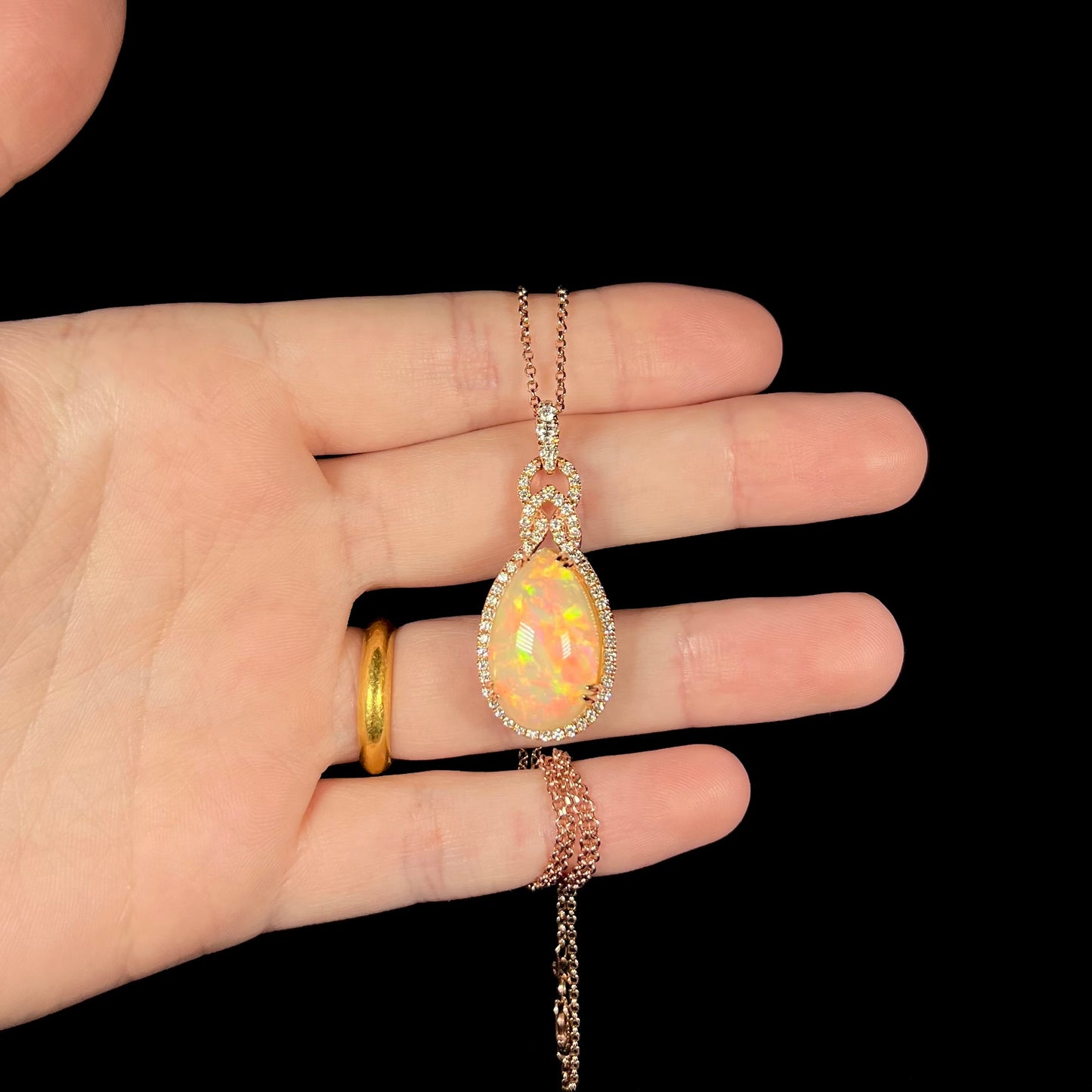 An pear shaped opal and diamond necklace made from rose gold shown held in a hand against a black background.