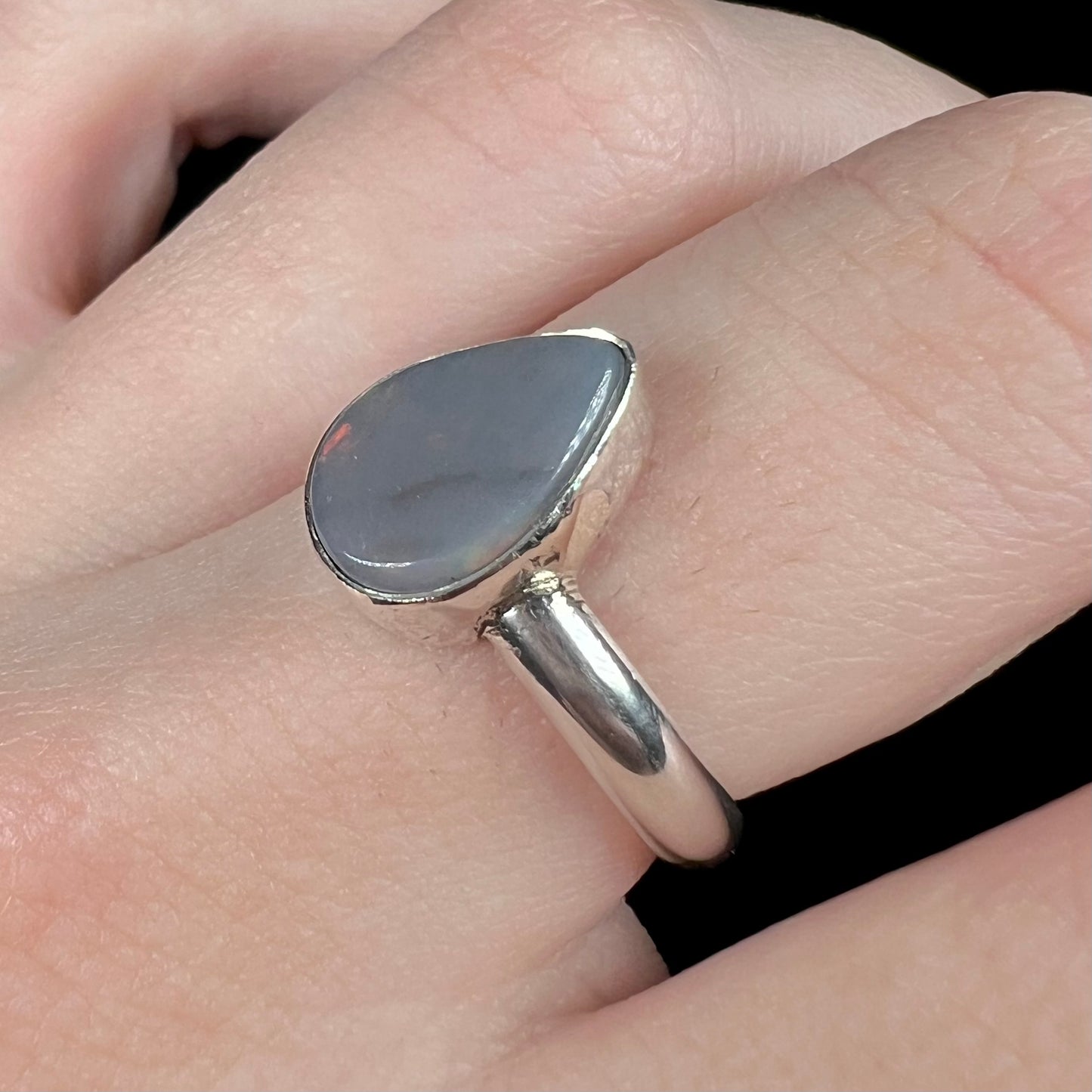 Angled macro of a pear shaped sterling silver dark opal ring worn on a finger.  The Australian opal's N6 dark body tone is apparent with no flashes of color.
