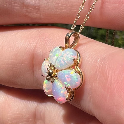 Close-up side view of a lab-created opal flower pendant in 14k gold, held in a hand under sunlight; the opals have a pink color