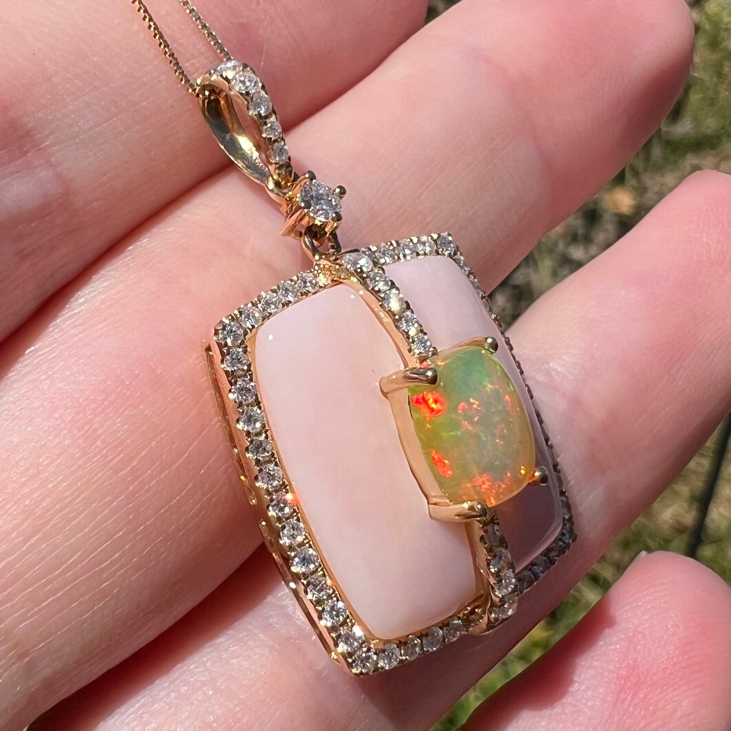 Angled close-up of a pink Peruvian opal and diamond necklace in 14k yellow gold with a fire opal accent, shown held in a hand in sunlight.