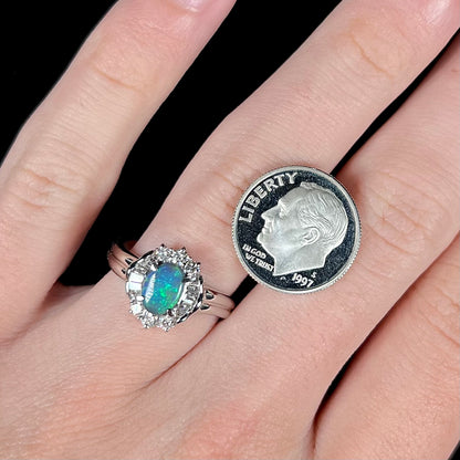 Top-down view of platinum black opal engagement ring worn on hand next to a US dime for scale.