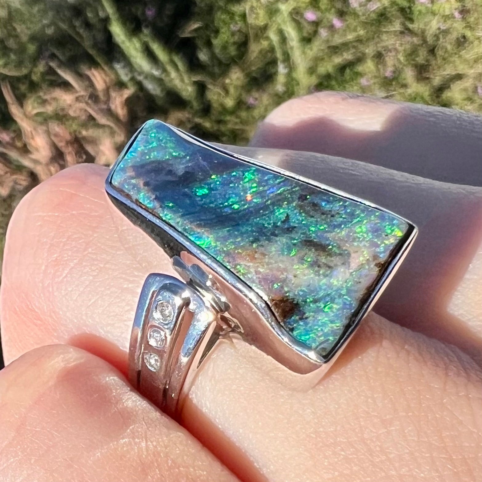 Angled sunlight view of a platinum boulder opal and diamond ring, worn on a hand against an outdoor background.