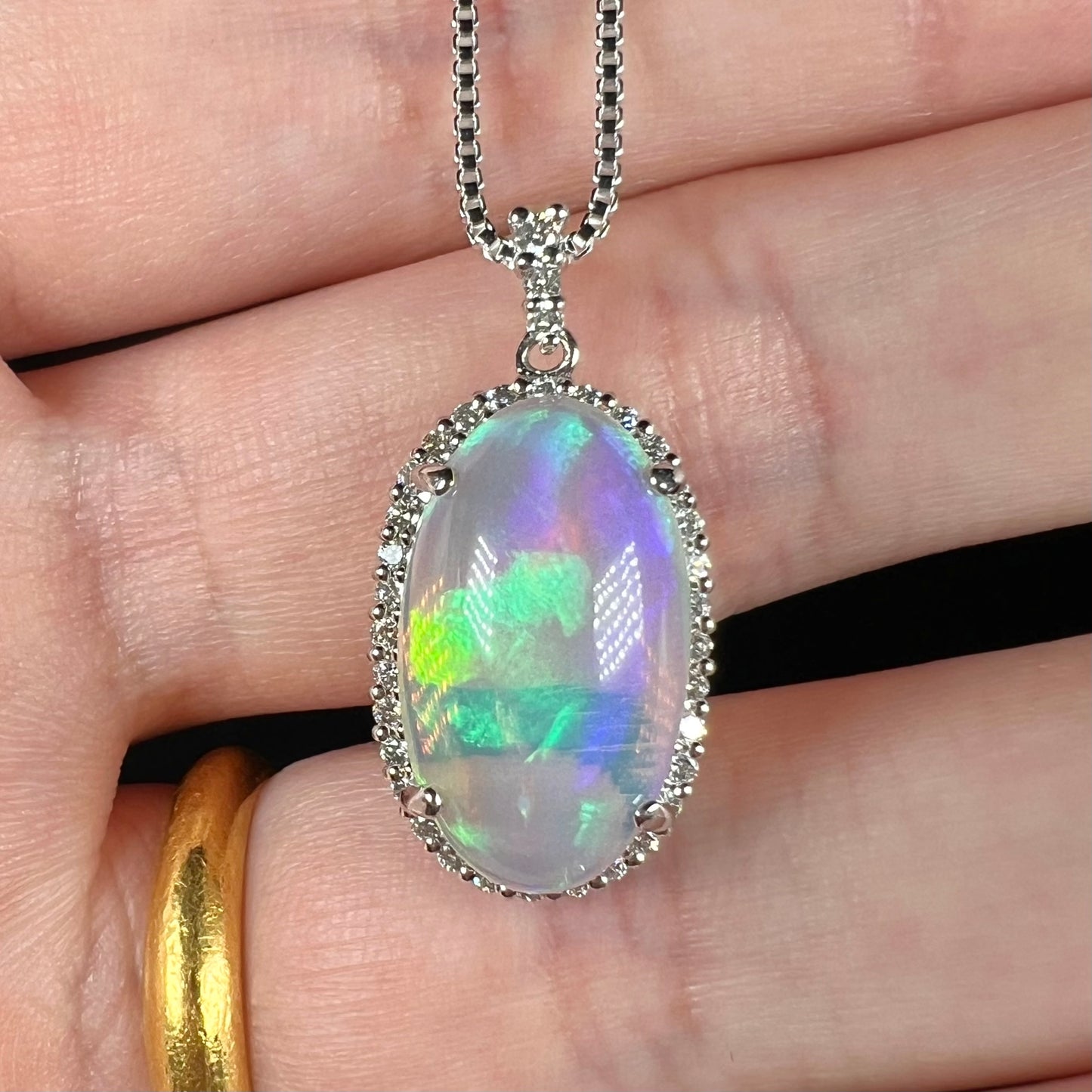 A platinum crystal opal and diamond halo pendant shown close-up in a hand.  The opal has subtle green and blue play of color.