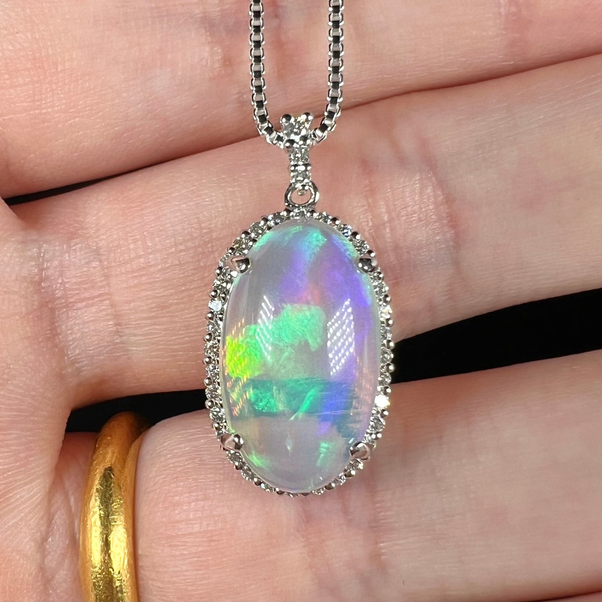 A platinum crystal opal and diamond halo pendant shown close-up in a hand.  The opal has subtle green and blue play of color.