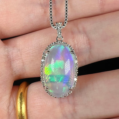 A platinum crystal opal and diamond halo pendant shown close-up in a hand.  The opal has subtle green and blue play of color.