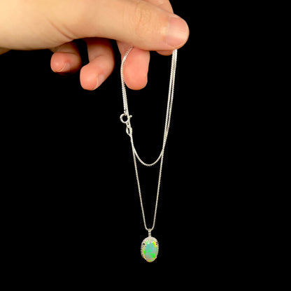 A platinum Ethiopian opal and diamond halo pendant, dangled from a hand by its silver chain against a black background.  The opal is green.