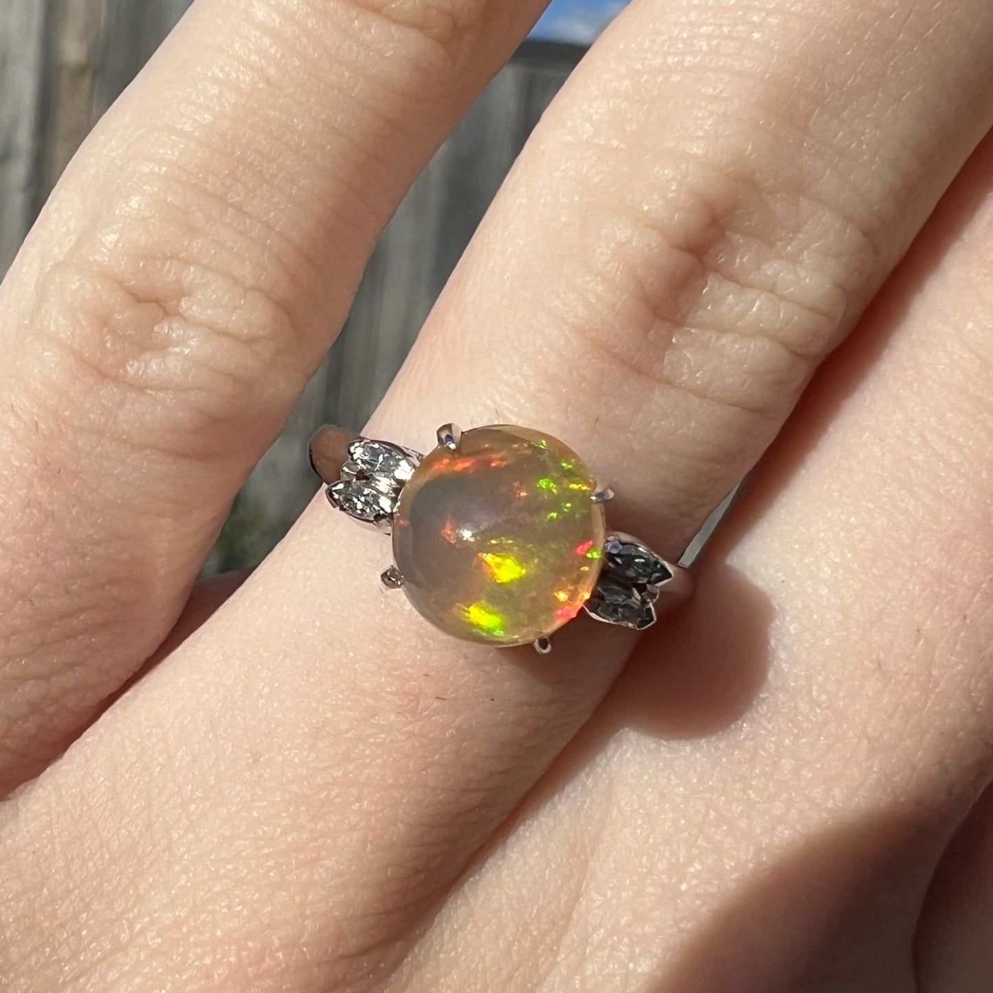 A platinum fire opal and diamond ring worn on a hand in the sunight.  Marquise cut diamonds accent the round Mexican fire opal.