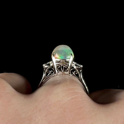 The gallery view of a platinum fire opal engagement ring worn on a finger, showcasing its Art Deco-inspired open filigree.