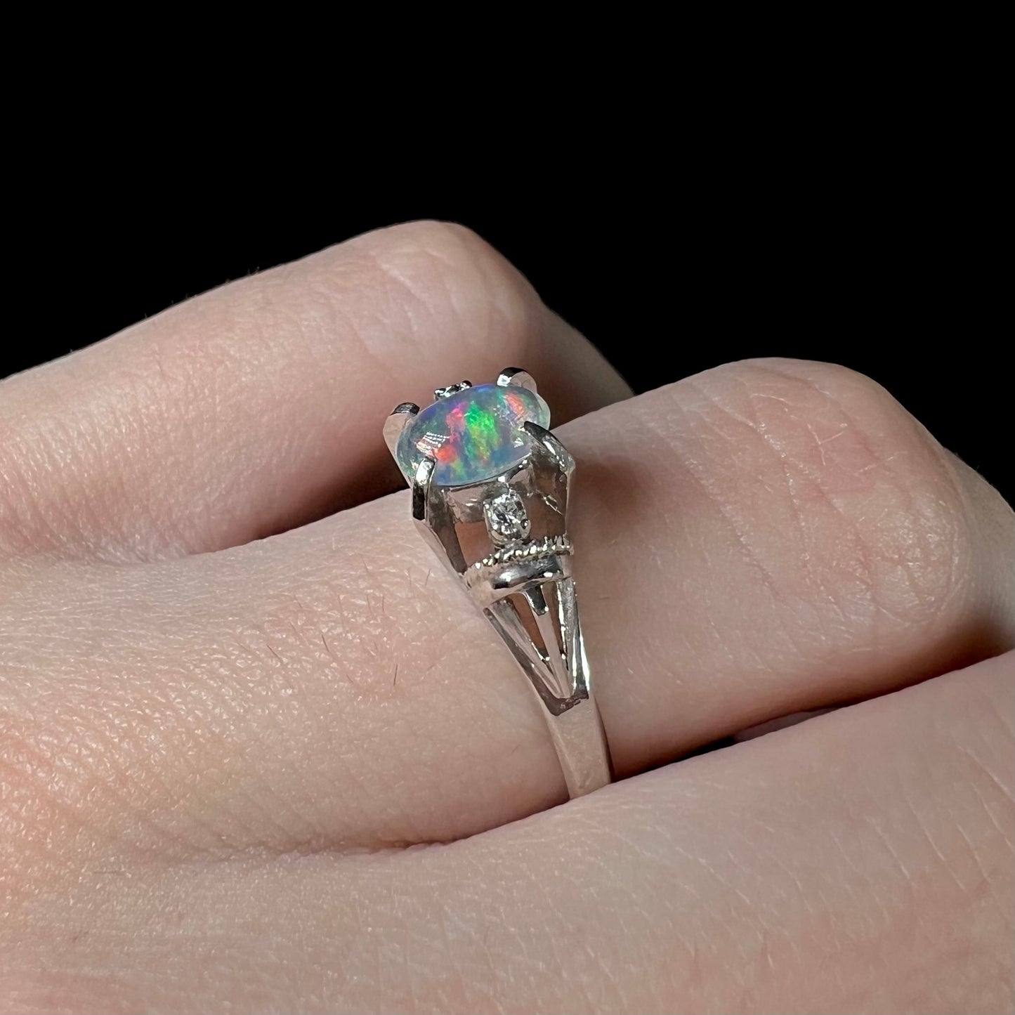 A green-red jelly opal and diamond engagement ring on a finger.  The ring has two diamond accents.