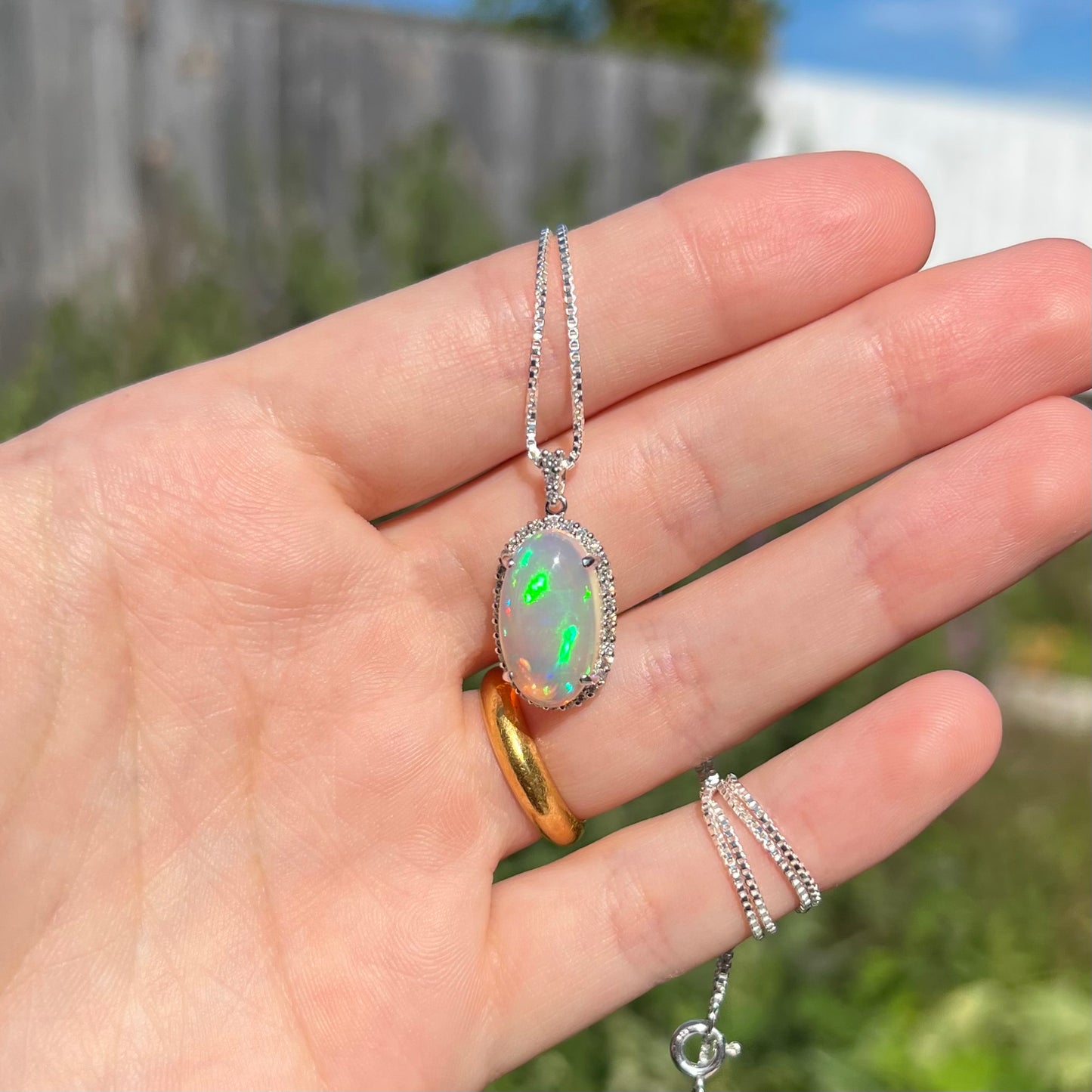 Hand holding a platinum crystal opal and diamond halo necklace in the sunlight against a blurred outdoor background.  The opal has green fire.