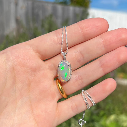Hand holding a platinum crystal opal and diamond halo necklace in the sunlight against a blurred outdoor background.  The opal has green fire.