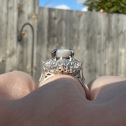 The gallery view of a vintage, Japanese-style platinum opal ring, worn on a hand in the sunlight.  Filigree and diamond accents are seen.