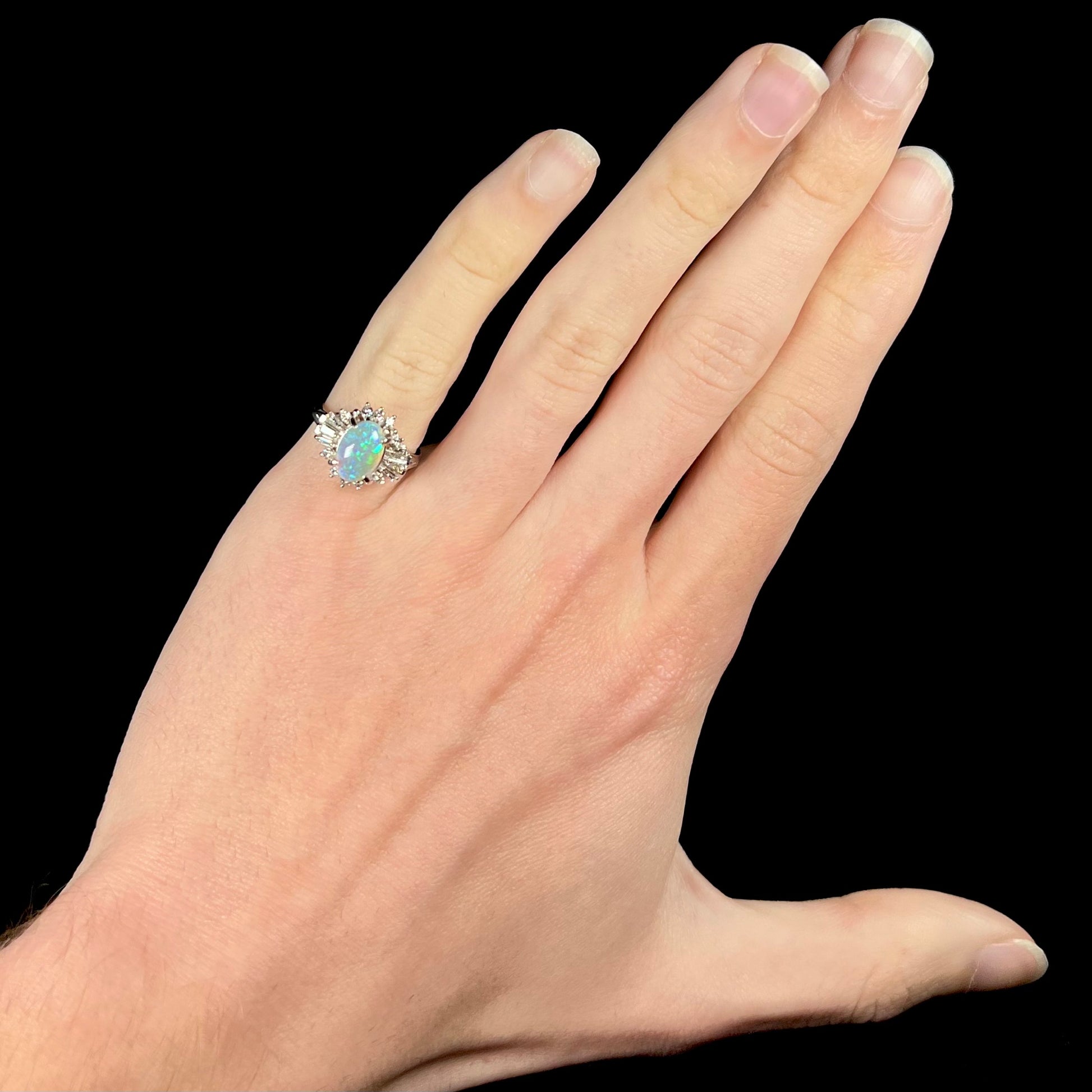 Hand wearing platinum semi-crystal opal engagement ring on pinkie finger over black background.