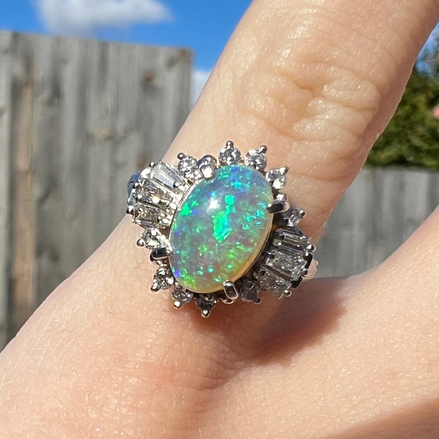 Macro closeup of platinum semi-crystal opal engagement ring worn on pinkie outside in sunlight, showcasing blue-green fire in the opal.