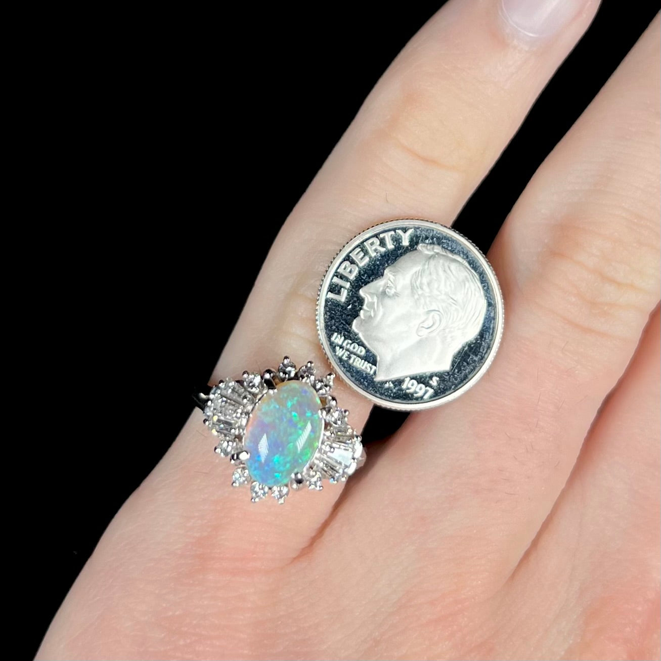 Platinum semi-crystal opal engagement ring worn on pinkie finger next to a US dime coin for scale, with black background.