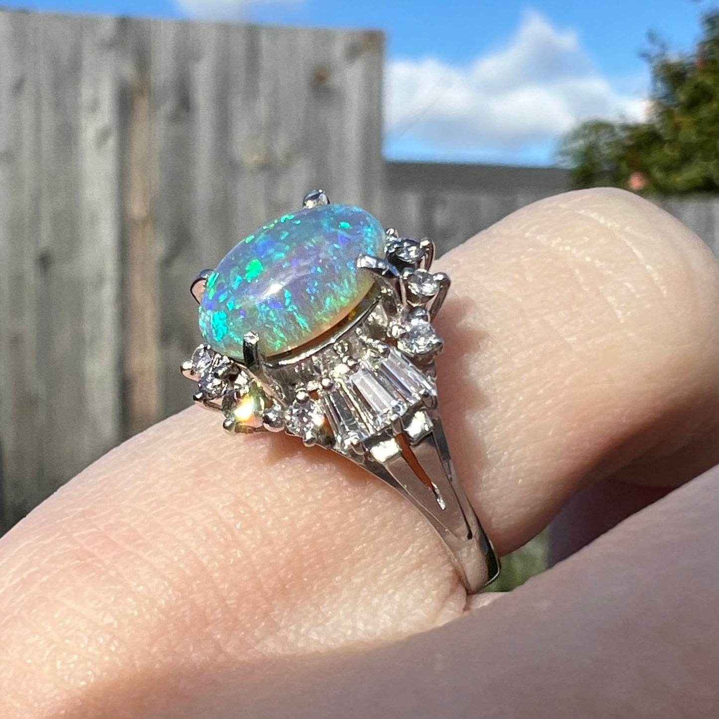 Diagonal angled macro view of platinum semi-crystal opal engagement ring on pinkie in sunlight, showing blue opal and baguette diamonds.