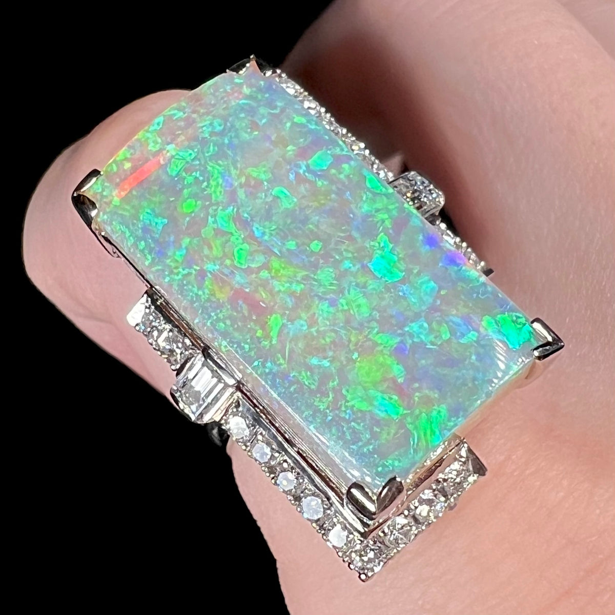 A mid-century Japanese cocktail ring set with a portrait-like rectangular Australian opal, framed by diamond accents, viewed from angle on finger.  The semi-crystal opal's body has pink reflections beneath green and teal play-of-color.