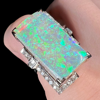 A mid-century Japanese cocktail ring set with a portrait-like rectangular Australian opal, framed by diamond accents, viewed from angle on finger.  The semi-crystal opal's body has pink reflections beneath green and teal play-of-color.