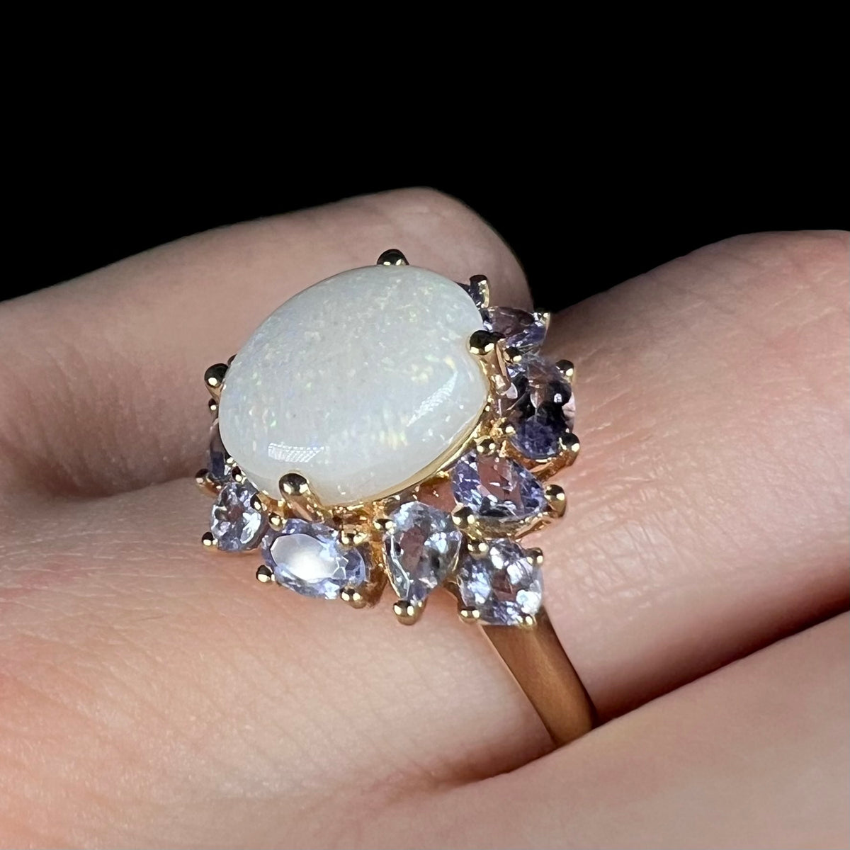 Angled close-up of prong-set Australian white opal ring with surrounding cluster of tanzanite accents, worn on finger; the ring's smooth yellow gold shank is in view