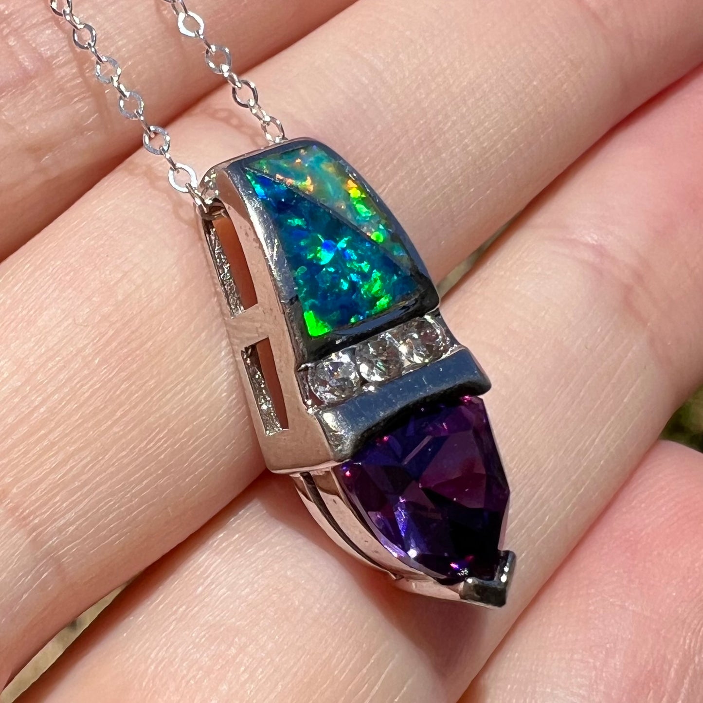 Angled sunlit close-up of a purple trillion cut cubic zirconia pendant made from sterling silver with lab-created opal inlay, resting against fingers.