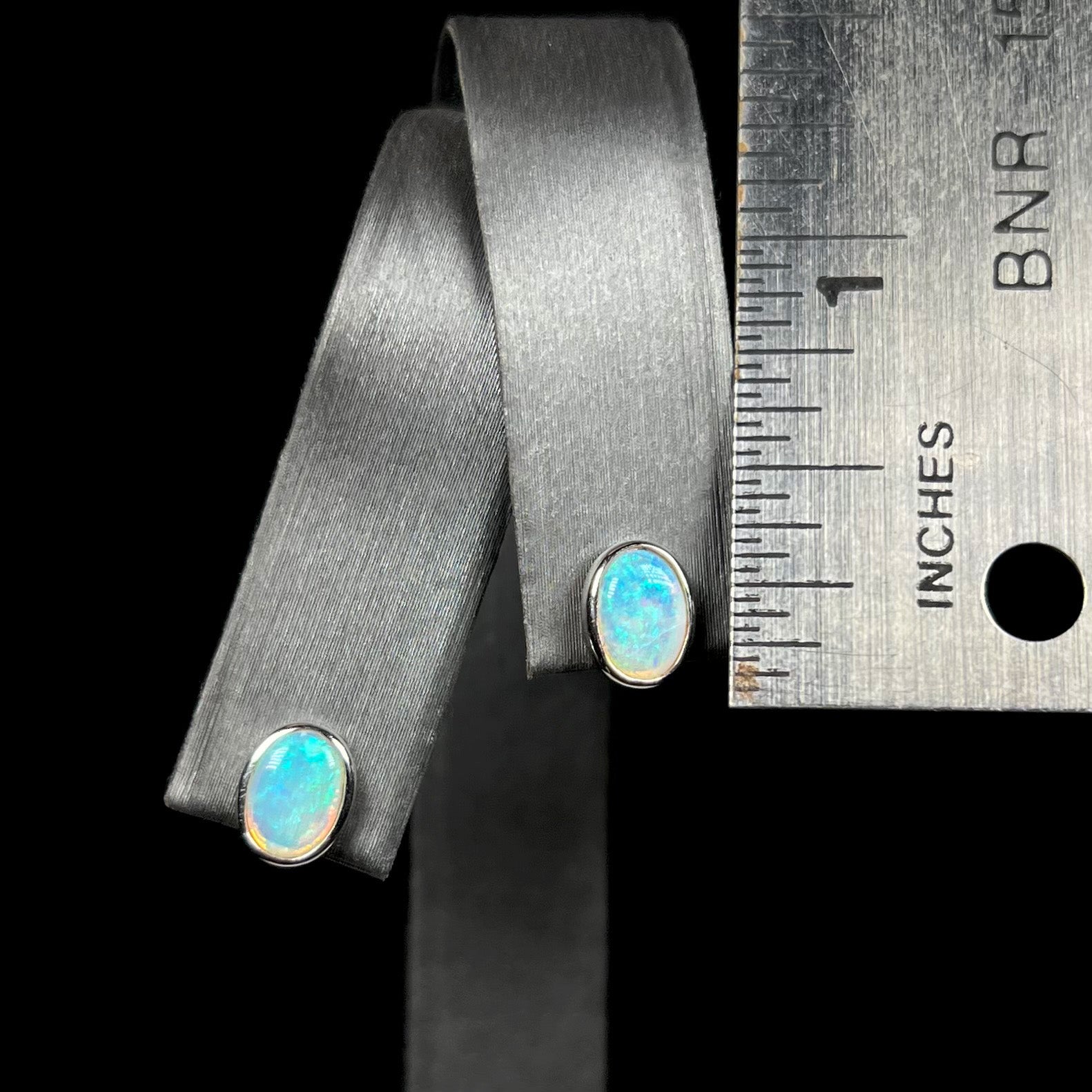 Dainty pair of white gold opal stud earrings shown on display next to ruler; the earrings measure approximately one quarter of an inch in length.