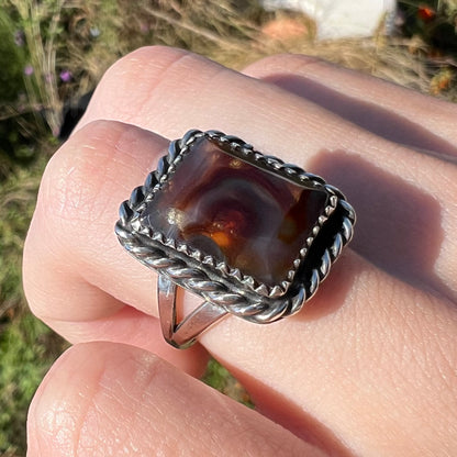 Rectangle cut fire agate ring with silver rope design and split shank, worn at an angle on a finger in natural light.