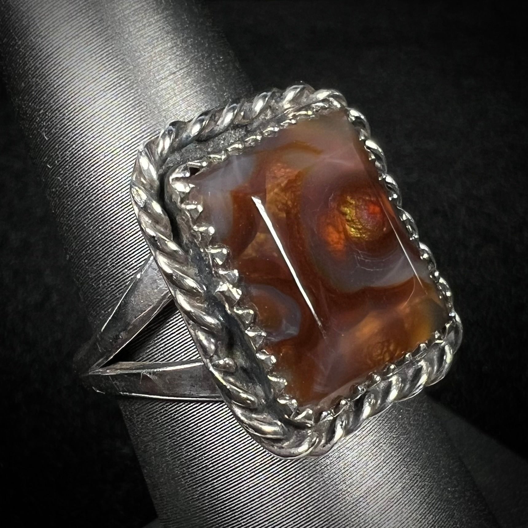 Angled view of sterling silver rectangle shaped fire agate ring with rope design against dark background.