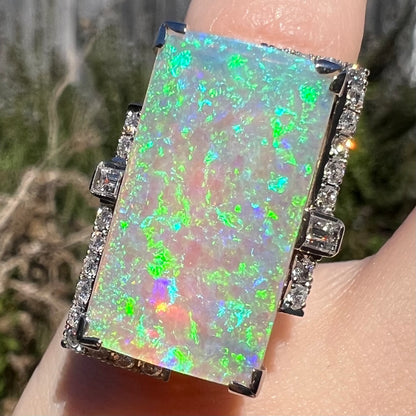 Close-up of a rectangular Australian light opal ring worn on finger in sunlight with brilliant green, blue, purple, and cyan play-of-color seen over smoky N7 body.