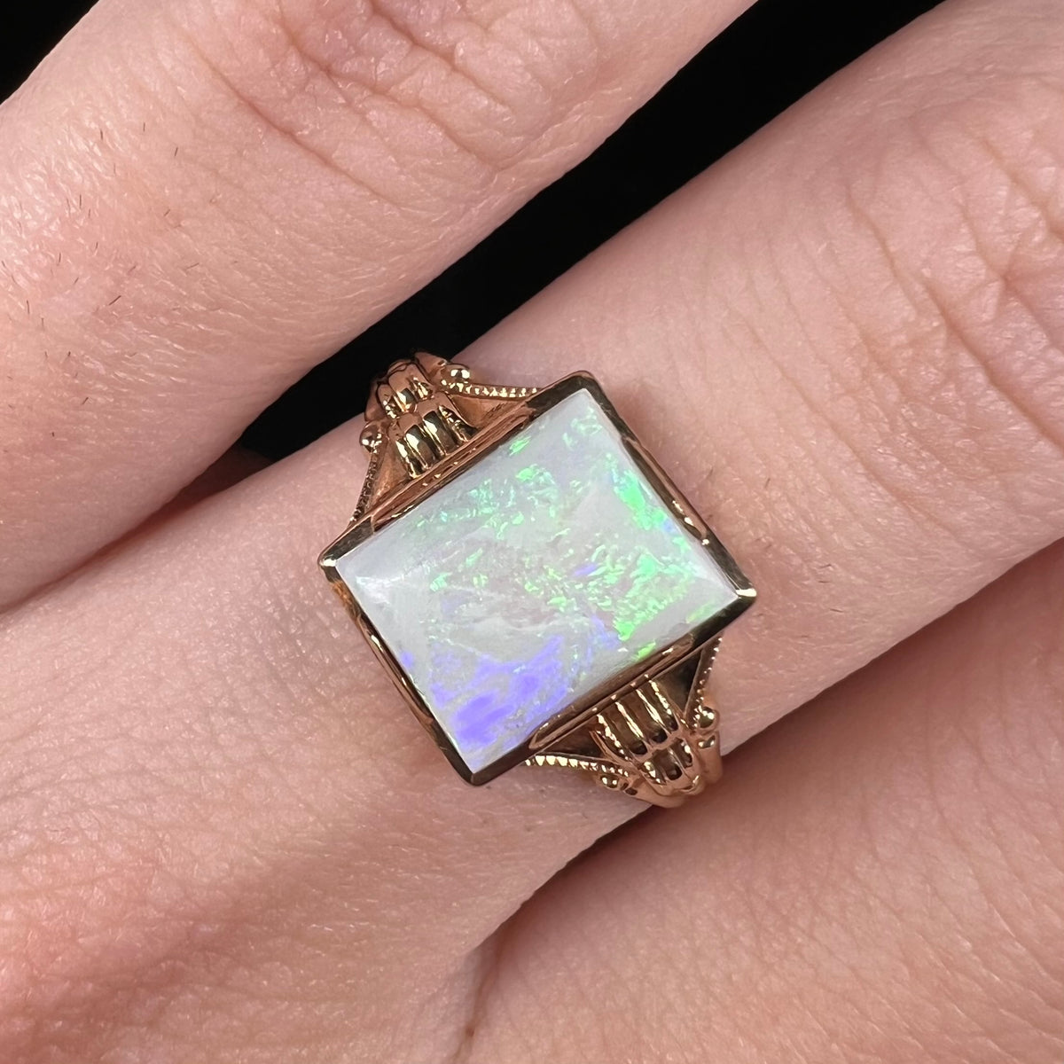 Close-up of rectangular shaped Australian opal solitaire ring with Art Deco-style design, worn on finger.  The opal has green and purple colors