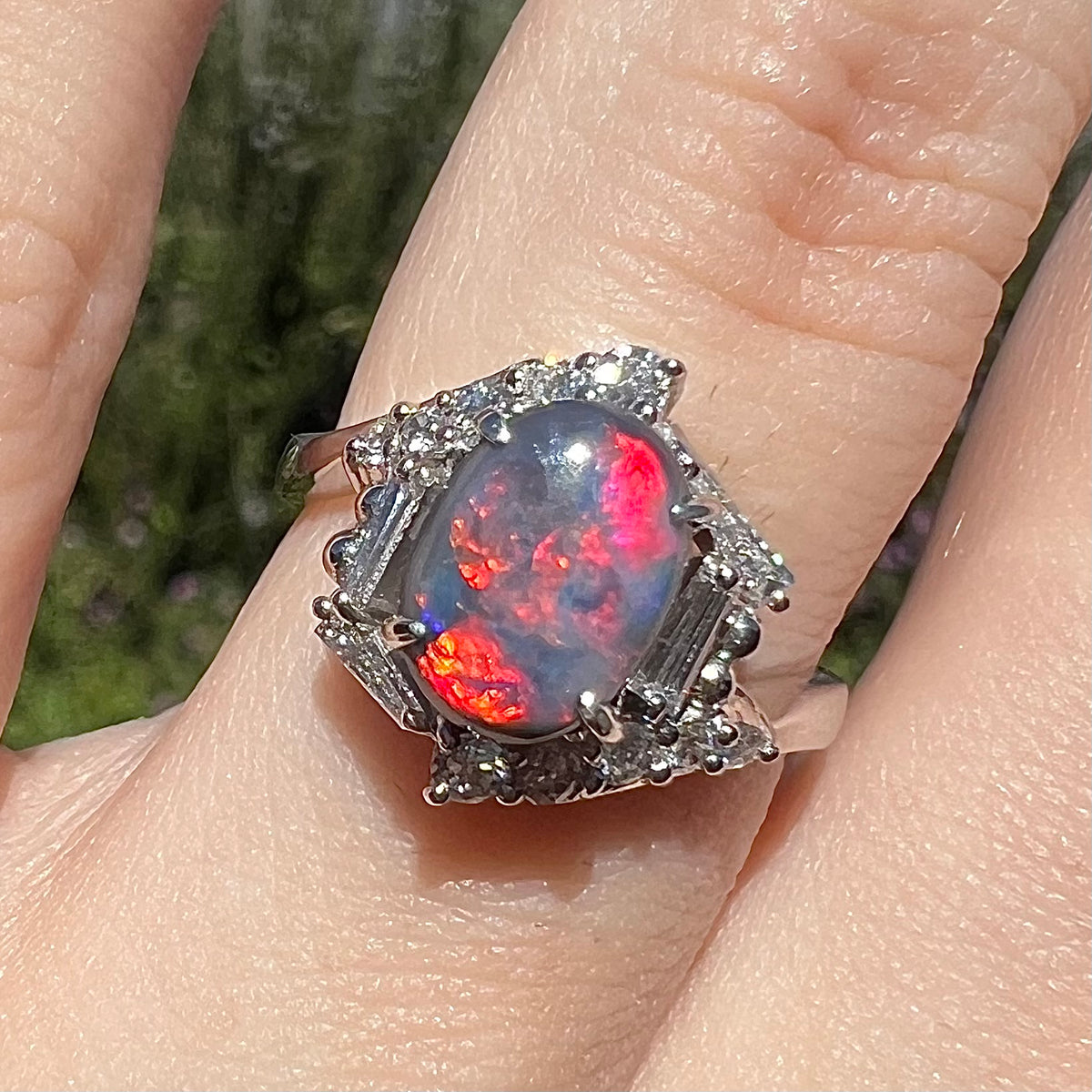 Close-up of vivid red and magenta-flashing Australian dark opal engagement ring, worn on finger in sunlight.  The ring has a hexagonal diamond halo design.
