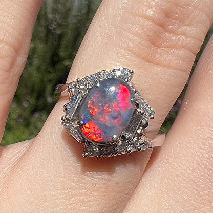 Close-up of vivid red and magenta-flashing Australian dark opal engagement ring, worn on finger in sunlight.  The ring has a hexagonal diamond halo design.