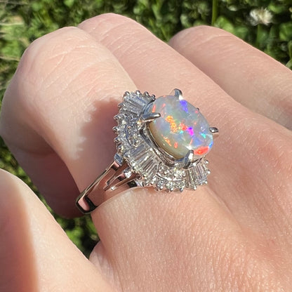 Close-up of a semi-black opal and diamond ring worn on a hand in natural sunlight, showcasing red and blue fire in a vintage platinum setting.