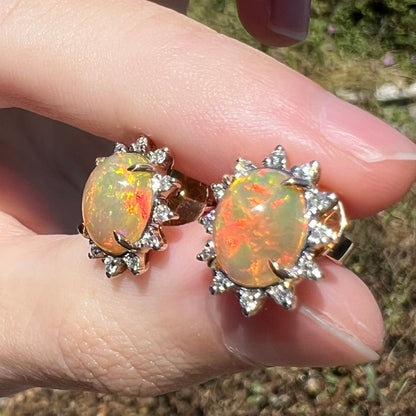 Red-playing Ethiopian opal and diamond earrings in 14k gold, held between fingers under natural sunlight