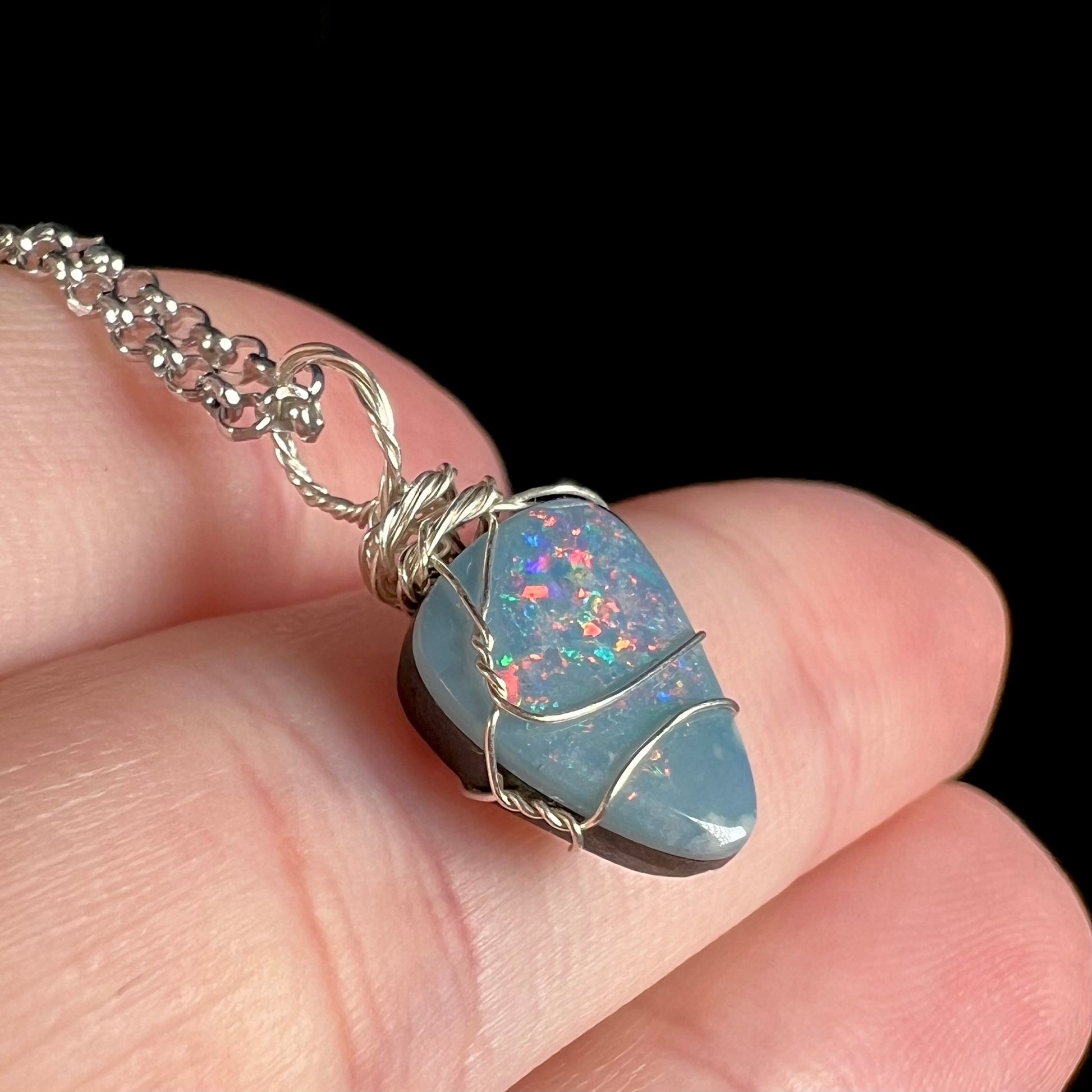 Angled macro close-up of red flashes in a freeform-shaped opal doublet mounted in a sterling silver wire wrapped pendant, held on top of fingers.