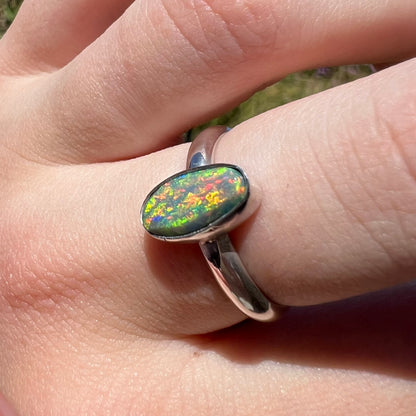 Angled close-up macro view of a red-multicolor Lightning Ridge dark opal women's ring in sterling silver, worn on a finger in natural sunlight.