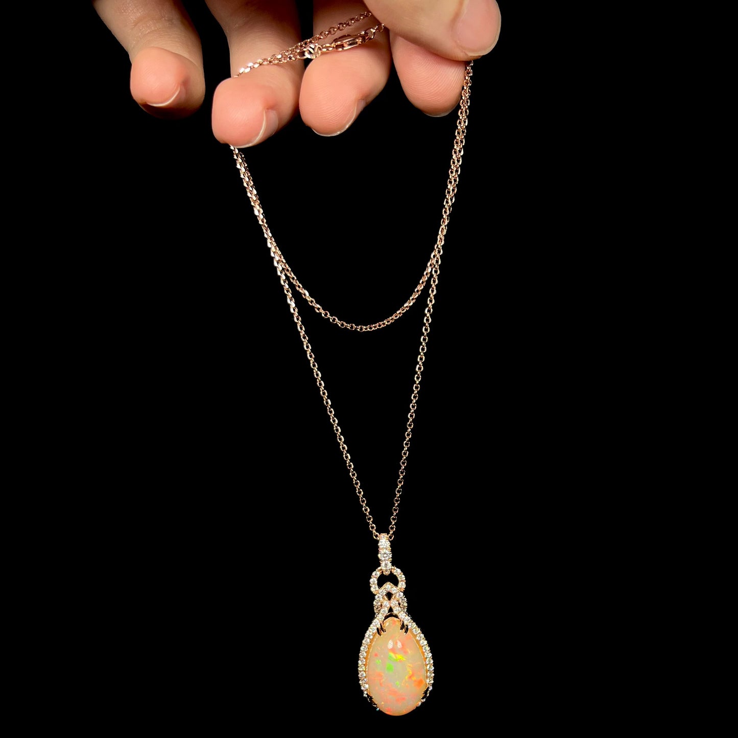 Hand dangling a rose gold pear shaped Ethiopian opal and diamond necklace by Le Vian from its cable-style chain against a black background.